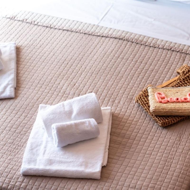 Letto ordinato con asciugamani bianchi e borsa estiva, atmosfera di relax e vacanza.